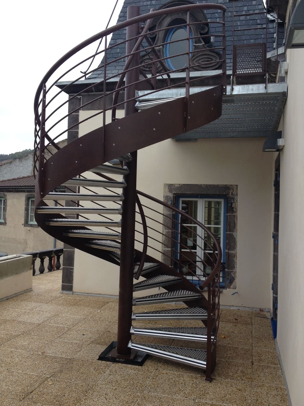 Un escalier en colimaçon extérieur avec une rampe en métal. L'escalier semble solide et bien entretenu, menant probablement à un étage supérieur d'un bâtiment.